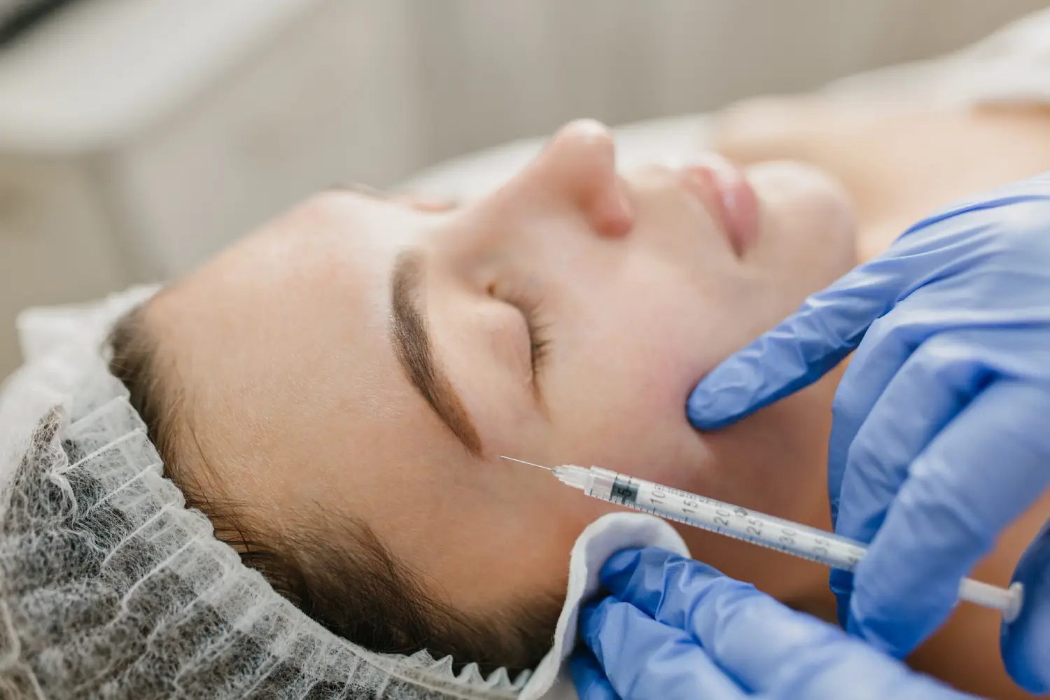 A close-up of a person receiving a skincare treatment, wearing a hairnet, with gloved hands administering a procedure.