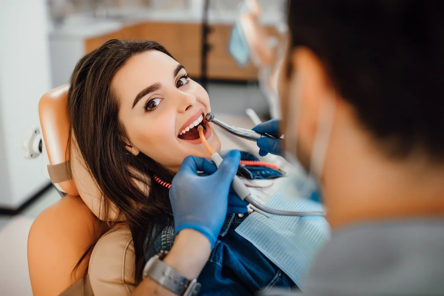 A dentist in gloves is using a dental tool while a patient reclines in a dental chair, surrounded by dental equipment.