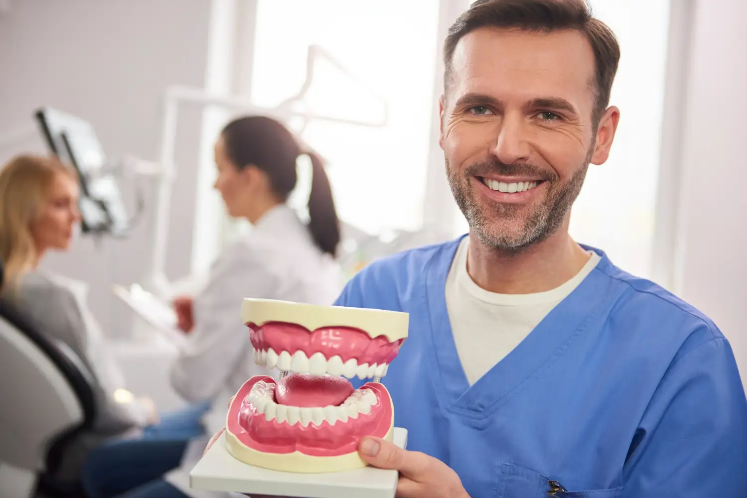 A dental professional in blue scrubs holds a large model of gums and teeth, while a patient is seated in the background.