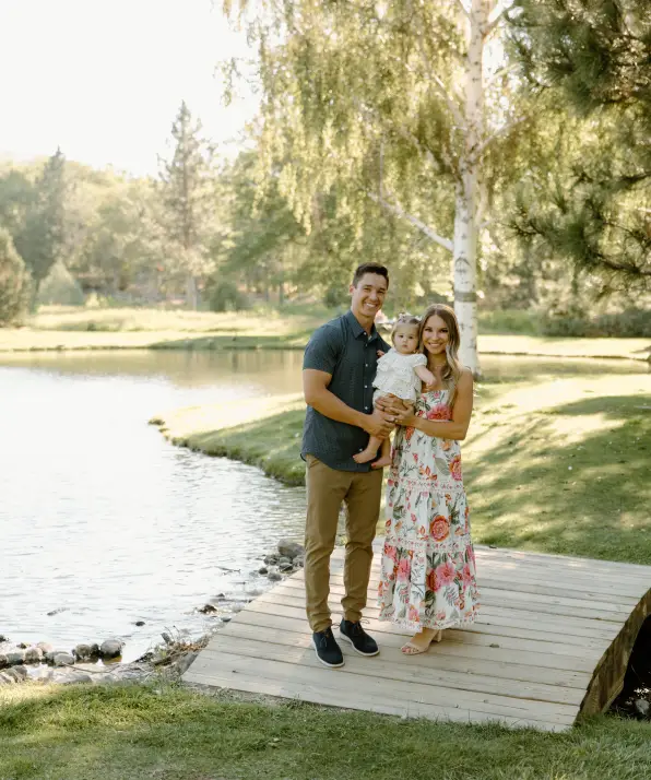 A family stands on a wooden dock by a serene pond, surrounded by lush greenery and soft sunlight, enjoying a peaceful moment together.