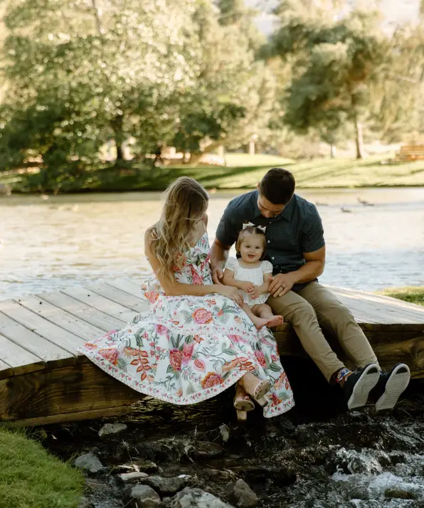 A family sits on a wooden dock by a serene lake, surrounded by lush greenery, enjoying a warm sunny day.