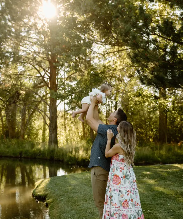 A joyful family scene by a pond, with a man lifting a child and a woman in a floral dress smiling nearby, surrounded by lush greenery.